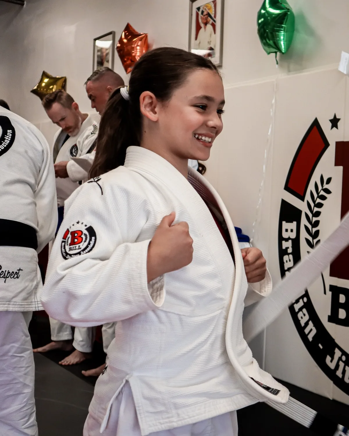 A smiling girl in a white gi getting her new belt tied on graduation day